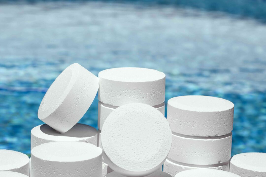 Photo of multiple white chlorine tablets stacked on top of one another. behind the tablets is a clear body of water that encompasses the whole background. 