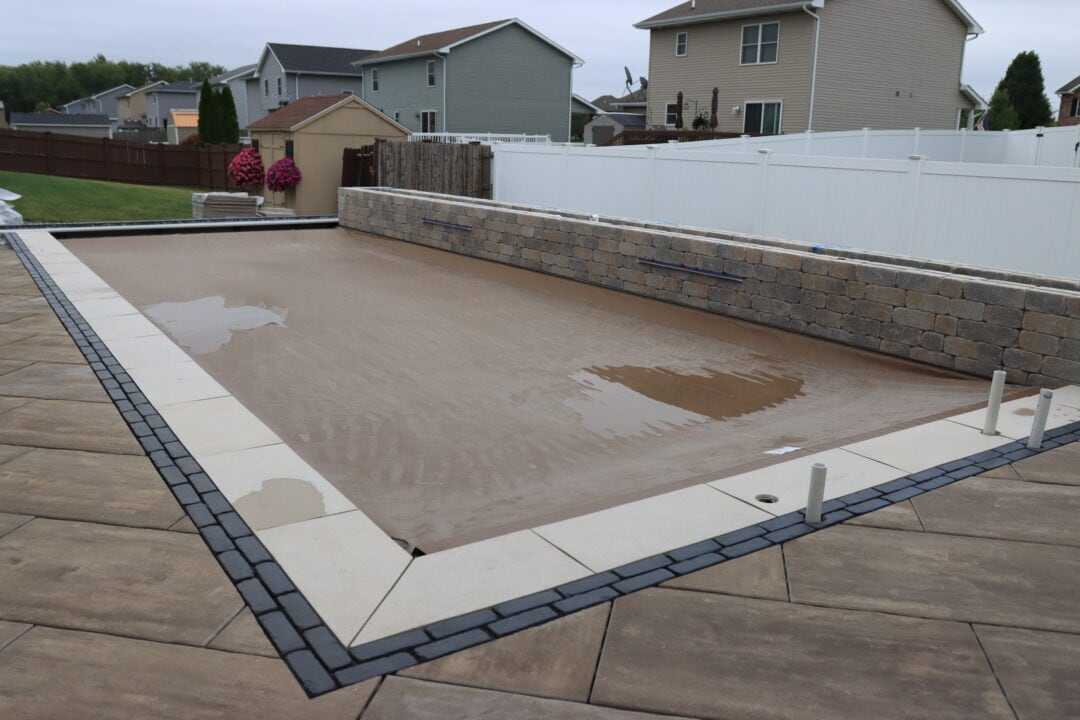 Closed inground pool covered by a brown automatic pool cover. The pool is surrounded by a concrete border, a black brick paver border, and a stamped concrete patio. Along the back-length of the pool is a retaining wall. A fence is behind the retaining wall. Houses can be seen in the background.