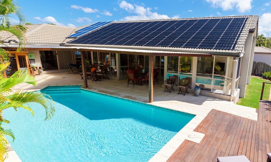 Photo of a backyard pool patio area. There is a large, light-blue inground pool in the foreground near a ranch-style house in the background. There are solar panels on the part of the roof closest to the pool.