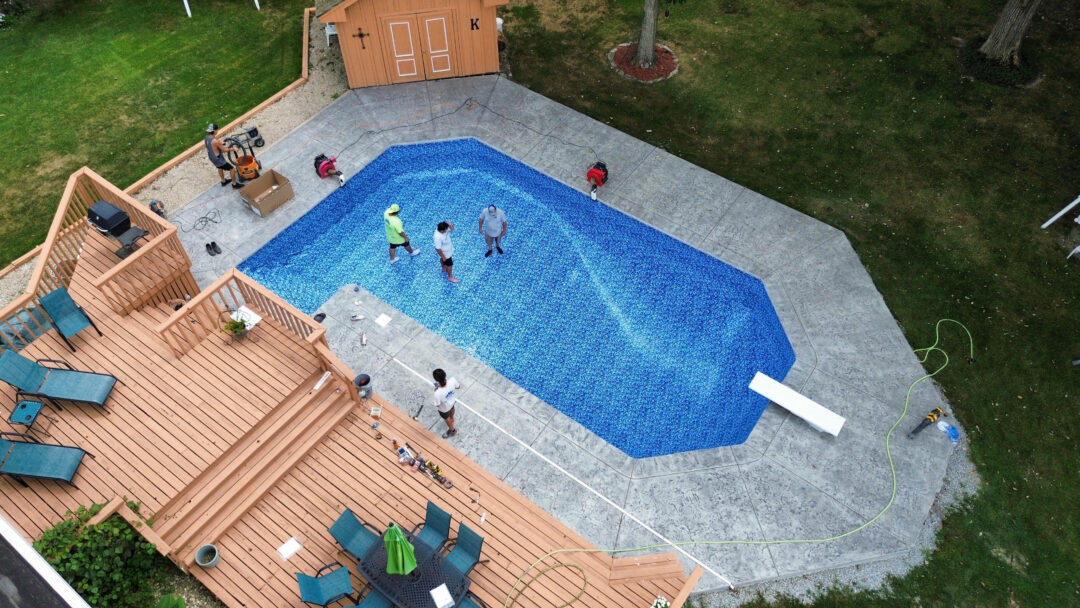 Aerial view of a backyard with an empty vinyl liner inground pool with a diving board surrounded by a large gray concrete patio. off the top left corner of the patio is a light-brown shed. Under the pool is a large light-brown wood deck with a black table and multiple blue chairs and tanning chairs. Three men stand in the shallow end of the pool and one stands on the patio near the middle of the wood deck.