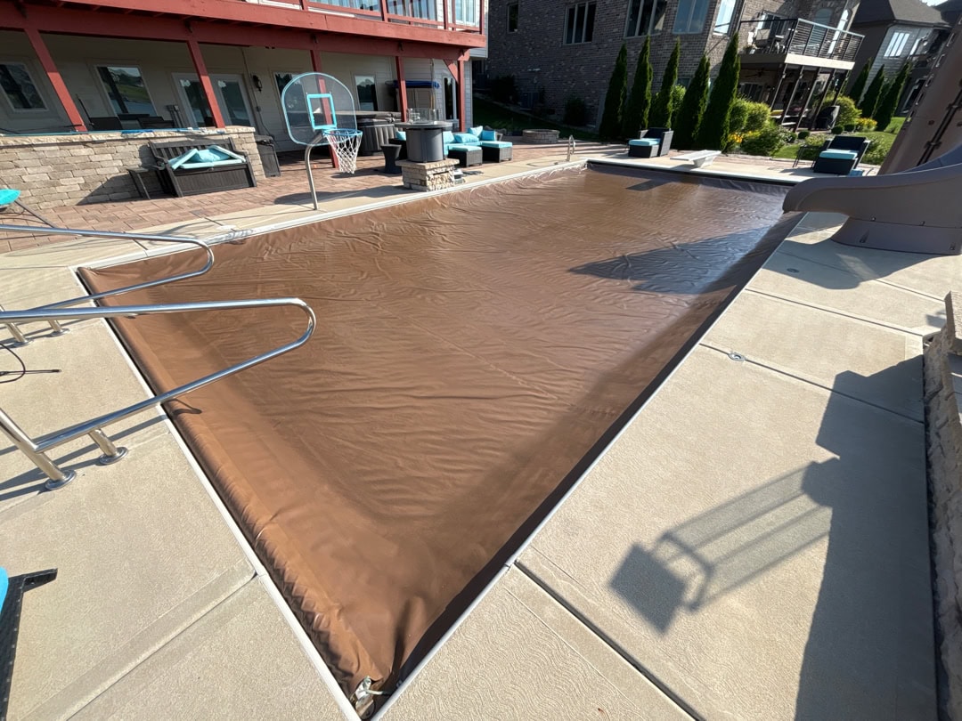 A wide shot of an inground pool with a slide, basketball hoop, and a clean, light-brown automatic pool cover.