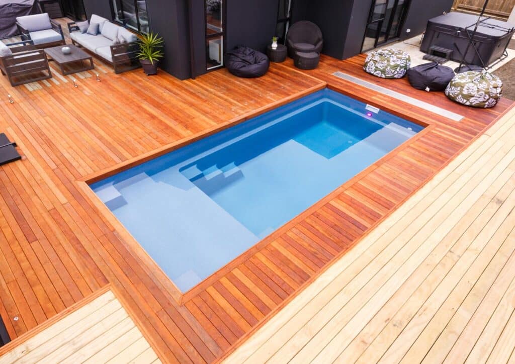 Landscape photo of a rectangular blue inground pool on a wood deck. The pool is surrounded by bright orange wood panels, atop which sit bean bag chairs and a table with chairs and a couch sitting against the house this patio is connected to. The house is barely visible at the top of the image. To the right of the pools deep end in the upper right corner of the image is a closed dark gray hot tub. 