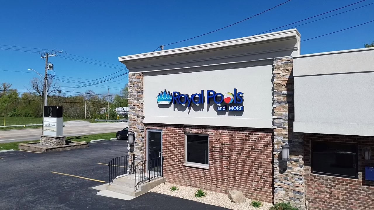 2025-05-09-15-04-33-755 Royal Pools and More retail store face. Logo is in front of tan wall with red bricks below, with one window in the middle and a door on the left.