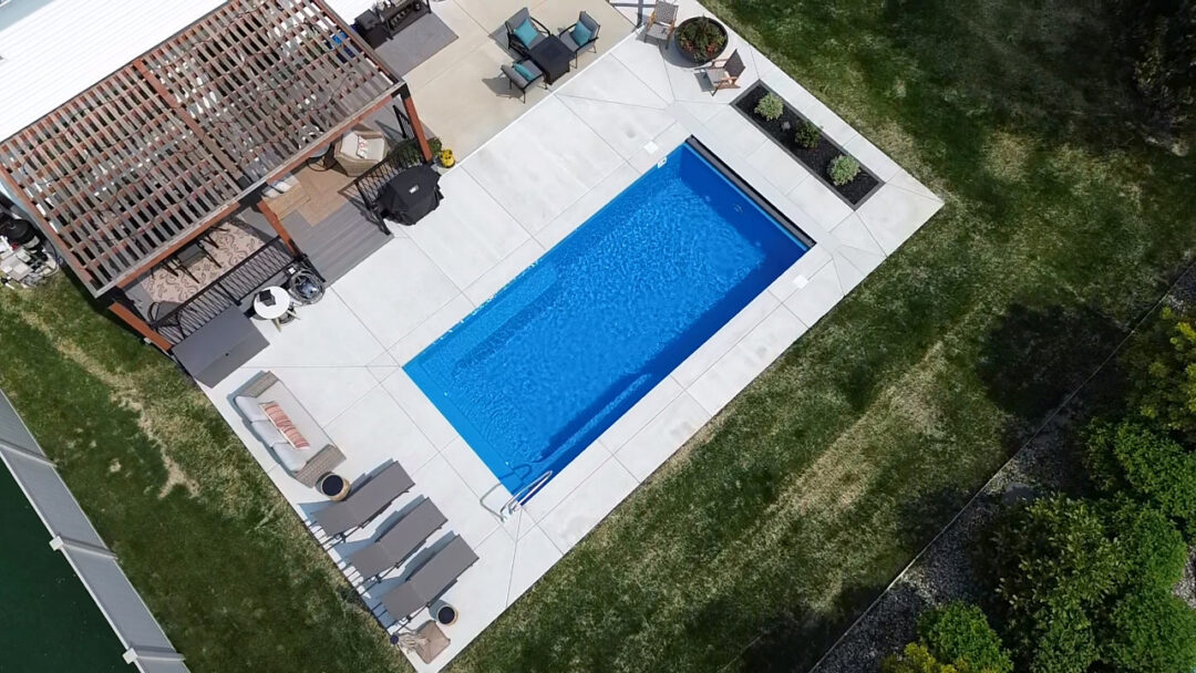 Landscape aerial photo of an inground fiberglass pool surrounded by light-gray concrete connected to the back of a house. Parallel to the corner of the house and pool patio is a brown pergola. Grass surrounds the patio on the edge of the bottom, left, and right side of the image. 