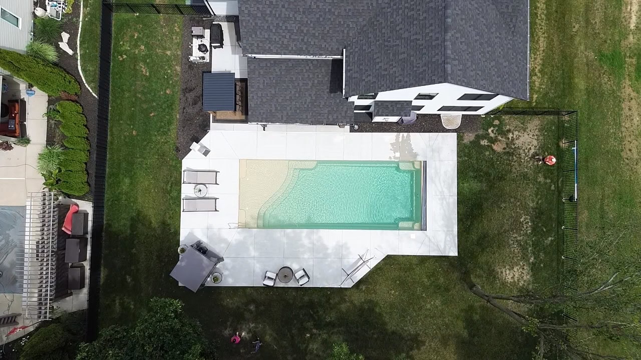 Aerial view of a sandstone rectangular inground pool surrounded by a concrete patio connected to the back of a white house with a dark-gray roof. Two chairs and a shade structure can be seen next to the shallow-end of the pool.