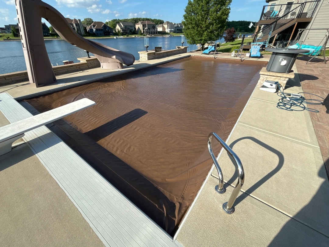 A shot of an inground pool on a raised deck with a brown automatic pool cover, slide, fireplace, and handrails overlooking a pond.