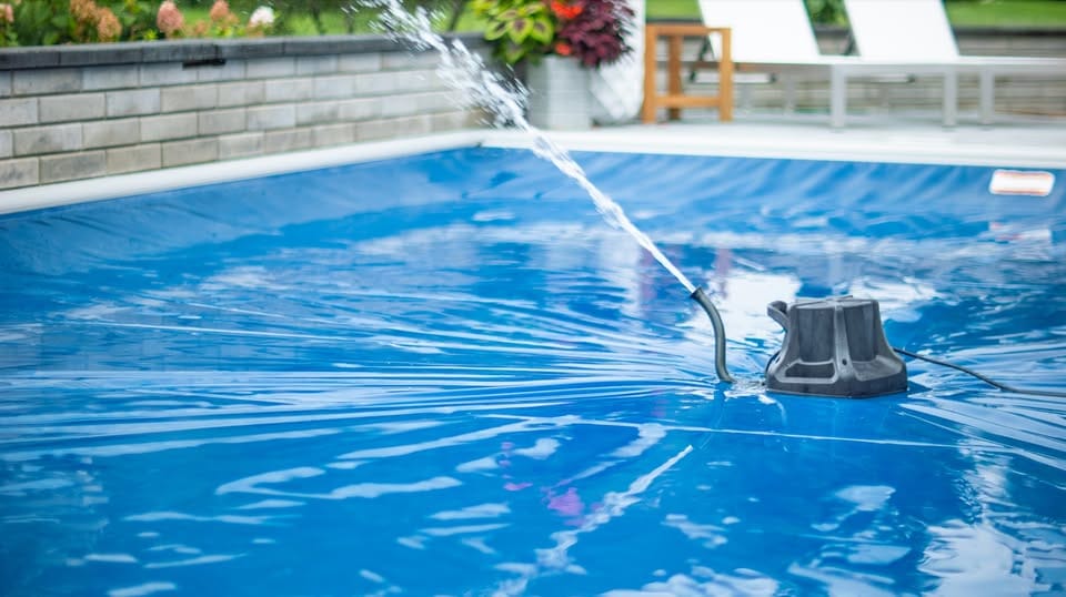 Landscape photo of a pool cover pump sitting atop an automatic swimming pool cover spewing water out of a spigot. The cover takes up a majority of the photo. a retaining wall can be seen surrounding the outside of the pool across the top of the image. 