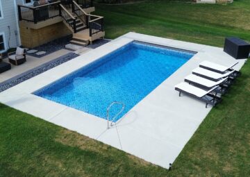 Photo of an inground vinyl liner pool surrounded by a concrete patio near a wood deck. Three tanning chairs are to the right of the pool on the concrete deck.