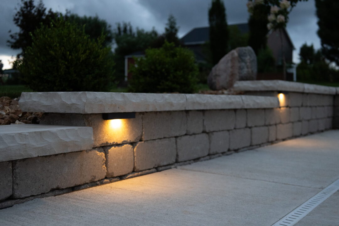 Landscape photo of a portion of a brick paver retaining wall featuring integrated ledge lights. The tops of trees and houses can be seen in the background in the top half of the image.
