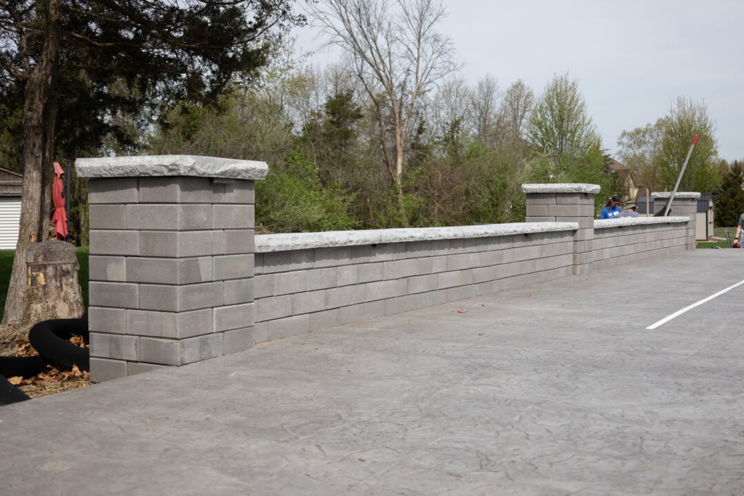 Landscape photo of a gray retaining wall propped up against a gray stamped concrete patio. The wall features three pillars, two on both ends and one in the middle. The retaining wall extends upward at an angle from the left side of the image to the right side. Trees can be seen in the background with the sky visible in the upper middle and right corner of the image.