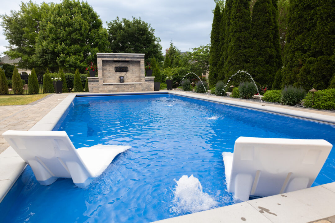 Landscape photo taken from in front of a blue inground fiberglass pool with a tanning ledge with bubblers. Two white lounge chairs sit on top of the tanning ledge in the foreground of the image. The pool is surrounded by a brick paver patio. On the left side of the image is a border garden running along the length of the pool, atop which sits multiple small ferns. in front of the garden, between it and the pool, two deck jets shoot arcs of water into the pool. In the background, parallel to the deep-end width of the pool, is a rectangular brick paver wall with a gap in the middle with wires visible. Behind the wall and around the pool are evergreen trees. Small evergreens line the yard behind the brick wall, and large evergreen trees line the side of the pool in the top right corner of the image.
