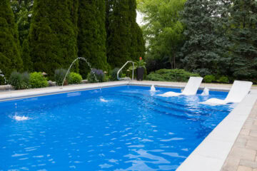 Landscape photo of a blue inground fiberglass pool surrounded by a white concrete border. The pool and surrounding patio takes up roughly the bottom half of the image. The top half of the image is comprised of surrounding evergreen trees and bushes. Three laminars are shooting arcs of water into the pool on the left/background side of the pool.