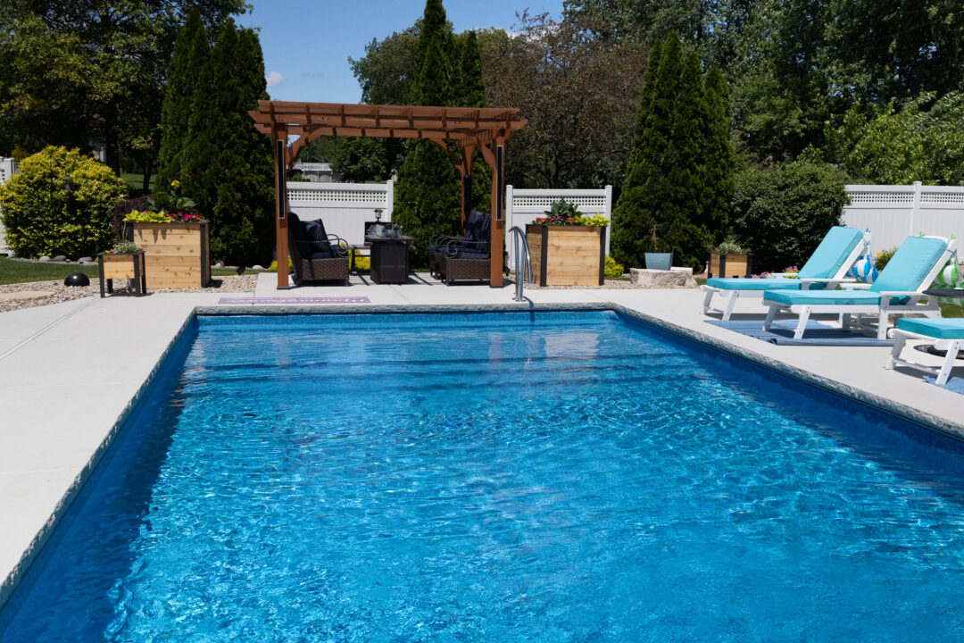 Landscape photo of part of a light-blue inground pool extending from the bottom of the frame just above the middle of the frame. The pool is surrounded by light-gray concrete on all sides. Light-blue lounge chairs can be seen cut-off by the frame on the right-side of the pool. A brown pergola sits even with the shallow end width of the pool near center frame. Bushes and plants line the frame on both sides of the pergola. Trees take up the rest of the background/top of the image.