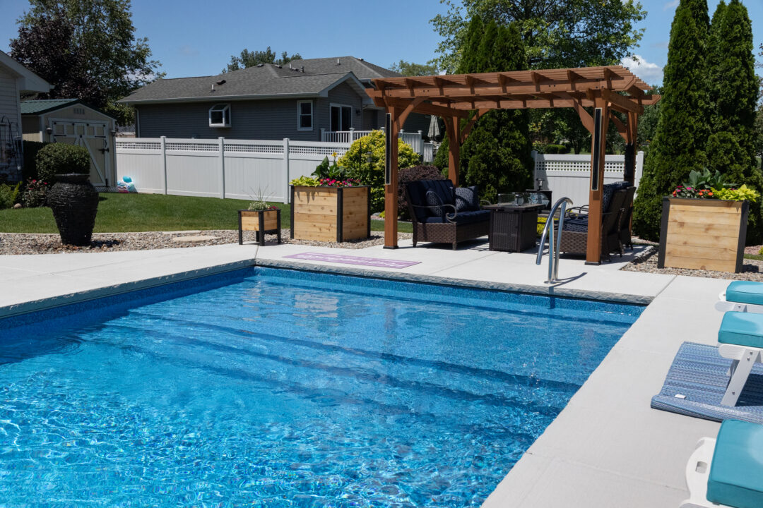 Landscape photo of part of a backyard featuring a blue inground vinyl liner pool surrounded by light-gray concrete. On the outside of the pool's shallow end is a brown pergola, under which sit chairs and a fireplace. Light-brown boxes sit on both sides of the pergola. Two large evergreen trees sit behind the pergola in front of a white fence. Houses and additional trees can be seen behind the fence in the background of the image.