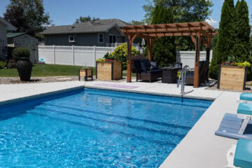 Landscape photo of part of a backyard featuring a blue inground vinyl liner pool surrounded by light-gray concrete. On the outside of the pool's shallow end is a brown pergola, under which sit chairs and a fireplace. Light-brown boxes sit on both sides of the pergola. Two large evergreen trees sit behind the pergola in front of a white fence. Houses and additional trees can be seen behind the fence in the background of the image.