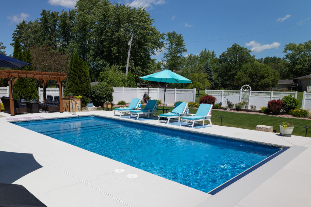 Landscape photo of a blue inground pool surrounded by gray concrete with grass in the background leading to a white fence. A pergola can be seen in front of the pool's shallow end on the far-left side of the image. Trees can be seen beyond the yard in the background.