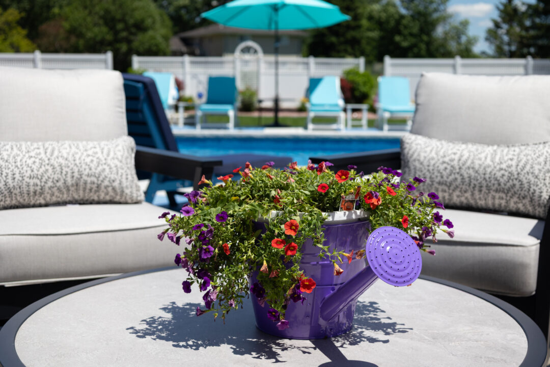 Photo of a potted plant sitting in a purple spout-pot atop a circular table. Two gray-cushioned chairs sit on both sides of the table. An inground pool and light-blue umbrella can be seen in the background directly behind the plant. Trees take up the top portion of the image spreading the entire length.