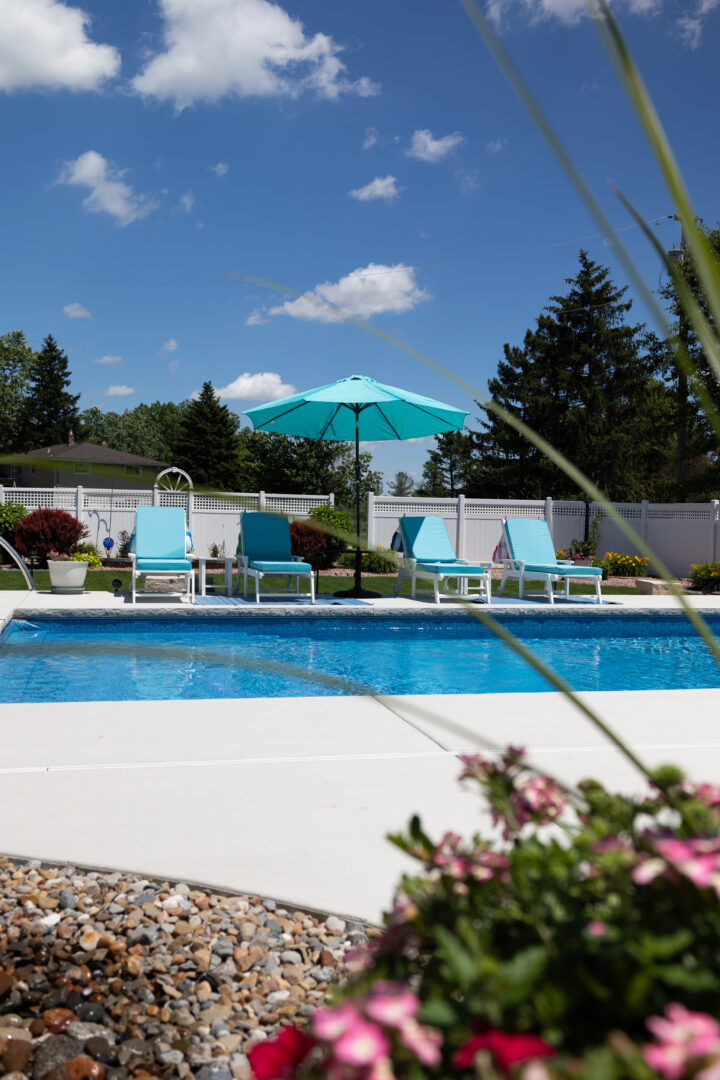 Portrait photo taken from behind a plant of an inground pool surrounded by a light-gray concrete patio, three light-blue lounge chairs, a light-blue umbrella, gravel which can be seen in the bottom left corner of the image. Treetops line the middle of the image stretching across the fence line. A blue sky with clouds takes up the upper-third of the image.