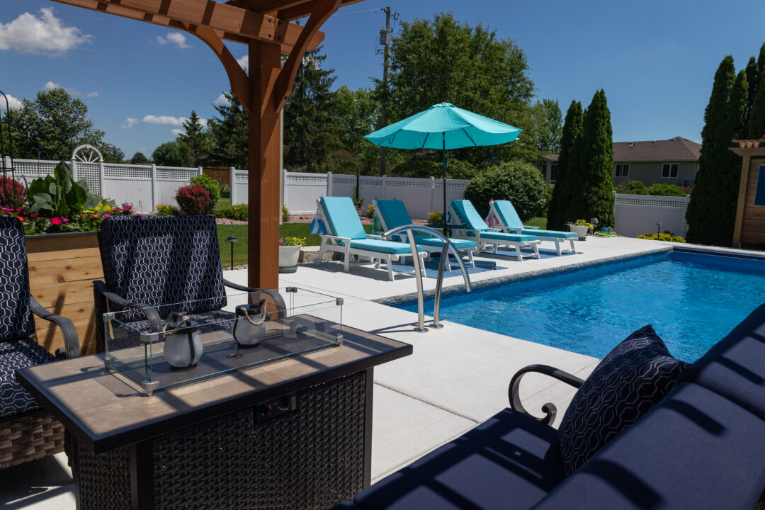 Landscape photo taken from under a pergola featuring chairs and a fire pit. Outside the pergola sits a light-blue inground pool cut off by the right-side of the image. On the left side of the pool, near center-frame are three light-blue lounge chairs and a light-blue umbrella. Evergreen trees sit in the front yard beyond the pool before a white fence. Treetops and a blue sky can be seen in the background beyond the fence.