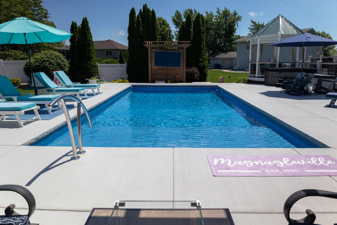 Landscape photo taken from in front of an inground pool surrounded by gray concrete and a white fence. On the left-side of the pool are three light-blue lounge chairs and a light-blue umbrella. Next to the chairs is a bush and two evergreen trees. More trees can be seen in front of the fence behind the pool. Houses and a blue sky can be seen beyond the fence in the background.