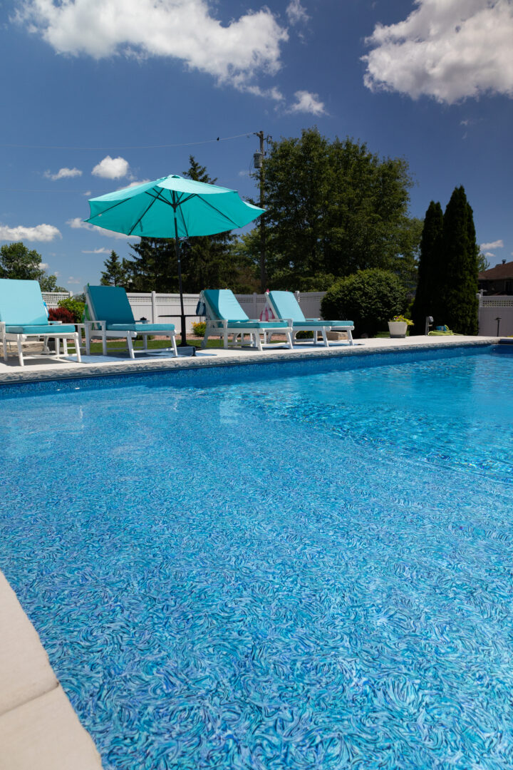 Portrait photo taken of a portion of an inground pool. The pool akes up most of the bottom half of the image. Concrete can be seen around the pool. A light-blue umbrella sits just outside of the pool on the patio surrounded by chairs. Evergreen trees sit in the yard in front of a fence in the background. Treetops and the blue sky can be seen in the background.