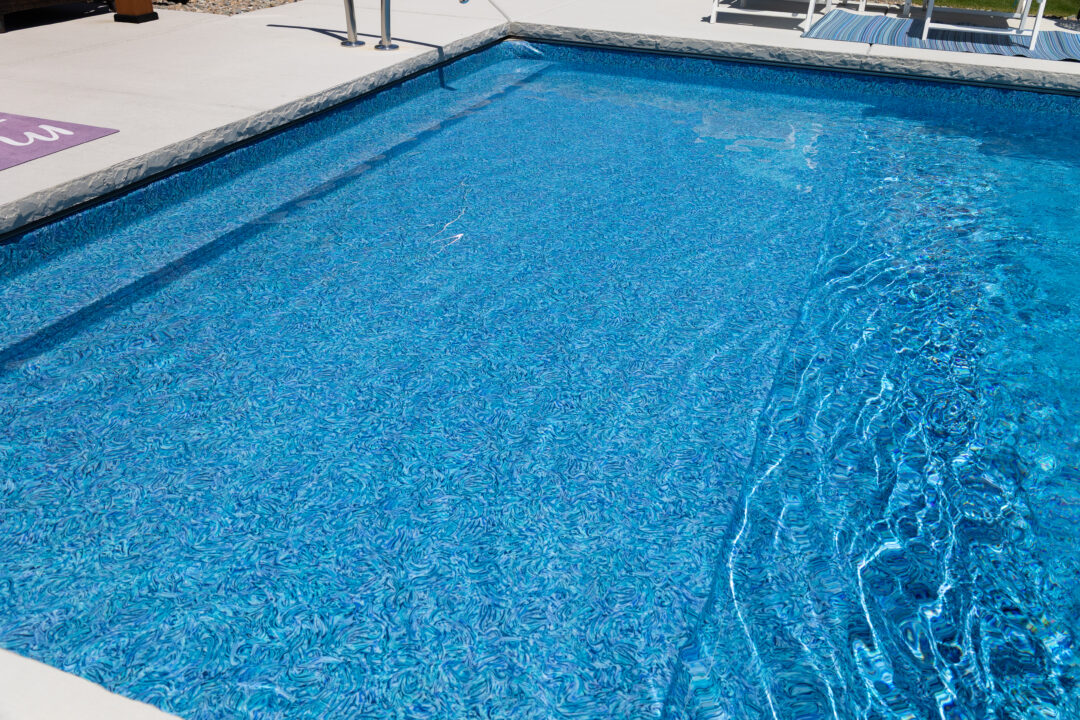 Landscape photo comprised primarily of part of an inground vinyl liner pool's shallow-end surrounded by light-gray concrete that can be seen on the top edge of the image and in the bottom left corner.