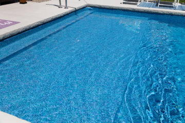 Landscape photo comprised primarily of part of an inground vinyl liner pool's shallow-end surrounded by light-gray concrete that can be seen on the top edge of the image and in the bottom left corner.