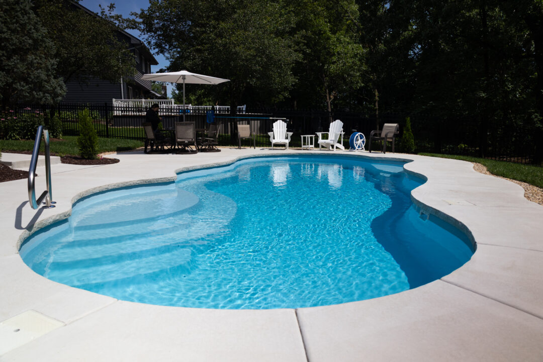 Landscape photo of a light-blue inground freeform fiberglass pool taken at a low angle in front of the shallow end. The pool is surrounded by light-gray concrete in the shape of the pool. Grass surrounds the patio in the background of the image. Tall trees and a black fence can also be seen in the background. Behind the pool's deep end are two white lounge chairs and a table with an umbrella and multiple chairs. A neighboring house and above ground pool can be seen beyond the black fence in the background. 