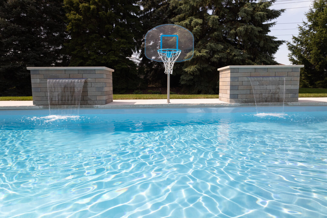 Landscape photo taken from one length-side of a light blue inground fiberglass pool. The pool and water takes up little over the bottom half of the image. Outside the pool are two rectangular spillover waterfalls placed equidistant from each side of the image spilling water into the pool. A basketball hoop with a light blue rim and target square sits in the middle of the image, between the two spillover waterfalls. Large evergreen trees can be seen in the background, beyond barely visible grass and concrete.