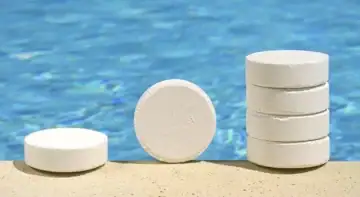 Centered image of six chlorine tablets at the edge of a concrete pool patio in front of the water. The tablet on the far left is lying down flat. The tablet in the middle is standing, and the four tablets on the right are stacked on top of one another.