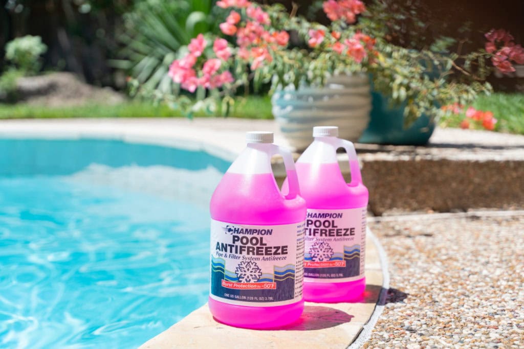 Two gallons of pool antifreeze sitting on a concrete deck surrounding an inground pool. Grass, shrubs, and potted plants can be seen in the background.