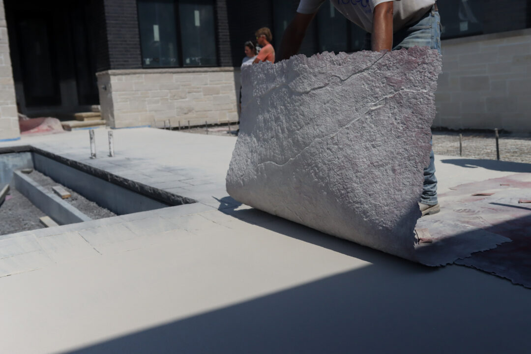Landscape photo of a concrete pool patio. A person is dropping down a square stamped pattern atop the concrete.