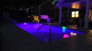 Inground pool with a purple pool light illuminating the water at nighttime. A glowing green and a glowing pink ball float in the pool. The pool features a slide on the left deep-end side, a basketball hoop, and a pool side gazebo. Next to the right-side deep end corner of the pool is an above ground hot tub also glowing purple.
