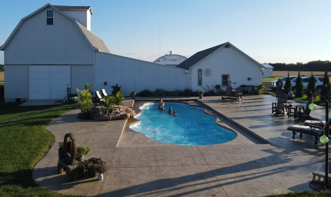 Landscape photo of a backyard comprised of a large stamped concrete patio. In the middle of the image is a light-blue, freeform inground pool with four people swimming in it. The pool is surrounded by multiple black-and-white lounge chairs. On the left side of the pool is a rock formation waterfall that spills over into the pool. Grass can be seen next to the patio on the far left side of the image. Behind the pool and patio is a large, white pole barn that extends across most of the background. An empty cornfield can be seen behind the barn. The sky is visible at the top of the image.