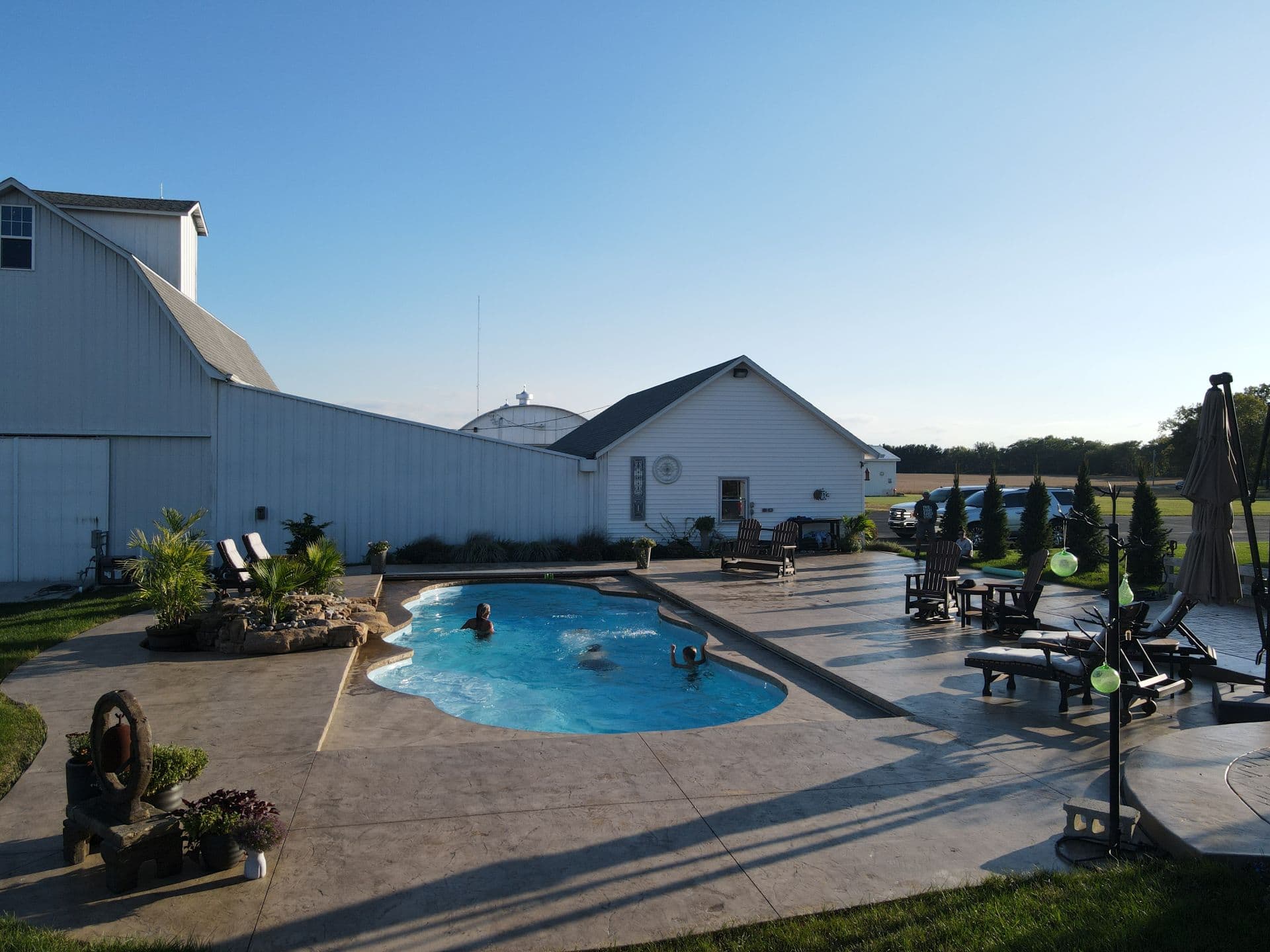 Photo of a light-blue freeform inground pool surrounded by a large stamped concrete patio. Off the left side of the pool is a rock-waterfall. Multiple chairs are on the deck around the pool. A row of trees can be seen on the far right along the edge of the patio. Behind the pool is a large white barn that stretches out left past the pool.