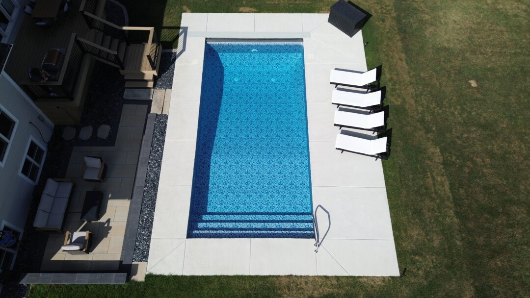 Aerial landscape photo taken from above an inground vinyl liner pool surrounded by a light gray concrete patio, atop which sit four black and white beach chairs parallel to the right side of the pool facing the grass, and a rectangular black container on the corner at the top of the image. Above, below, and to the right of the pool and patio is green grass extending to the edges of the image. To the left of the house is a smaller decking area and raised wood deck connected to a light-gray house at the ;eft side edge of the image.