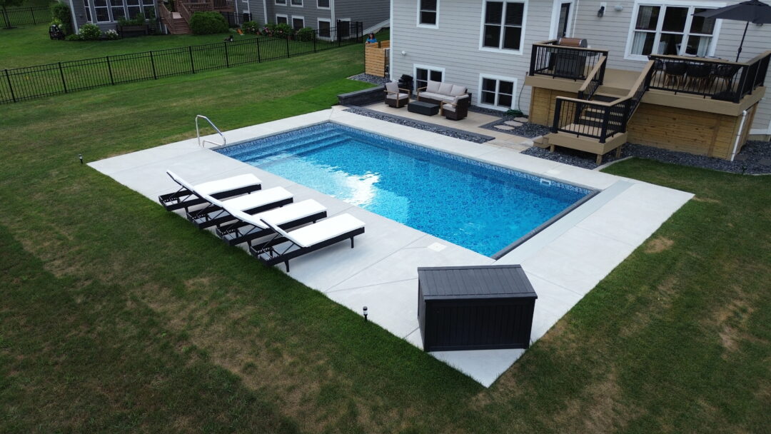 Aerial landscape photo of a backyard featuring an inground vinyl liner pool surrounded by a light-gray concrete patio. On the left-side shallow end corner of the pool is a handrail. Next to the pool between it and the grass surrounding the patio are four black and white beach chairs. In the corner closest to the bottom of the image is a large black container. The patio is connected to a smaller patio area connected to a light-gray house with a light-brown wood deck that descends down toward the pool near the top right corner of the image. In the background in the top left corner extending up to the middle of the image is a black fence connected to a neighboring house.