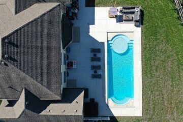 Aerial shot of a backyard and the back-half of a house with a large concrete patio encasing a light-blue roman-shaped inground pool.