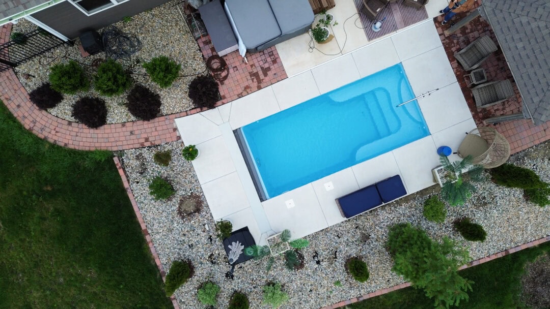 Aerial landscape image of a light-blue inground fiberglass pool surrounded by a light gray concrete patio, which is connected to the back of a house and surrounded by a rock bed on the outer edge. Small bushes and evergreen trees sit atop the rock bed. Outside the concrete and rock is grass in the bottom left and right corners of the photo. The top of a gazebo can be seen cut off by the edge of the top right corner of the image. 