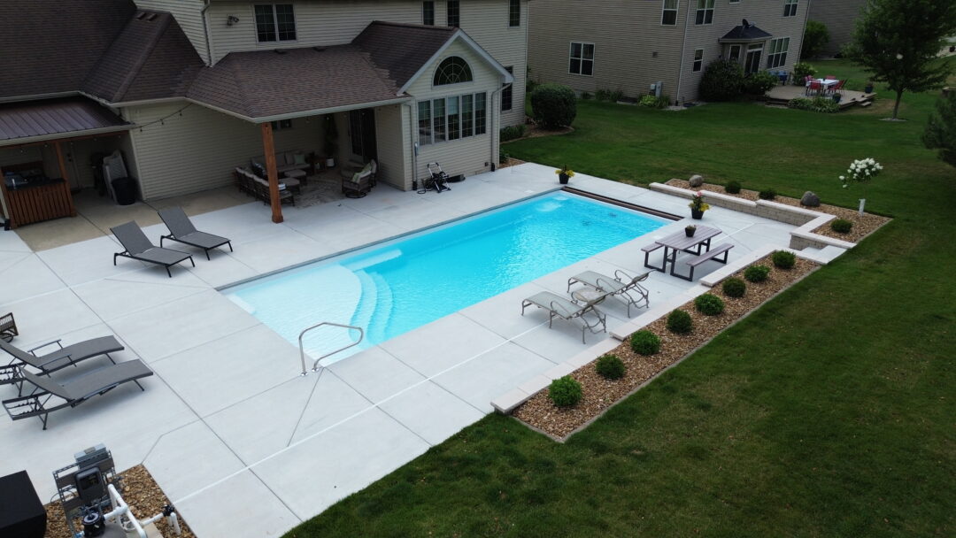 Landscape photo of a white fiberglass pool surrounded by a light-gray concrete patio connected to the back of a house. In the foreground of the image behind the patio is a long garden bed that wraps around the outer corner of the image. Lounge chairs rest on both length-sides of the pool. 