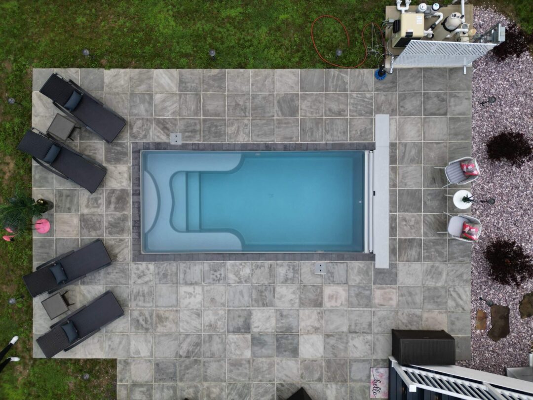 Aerial landscape photo of a portion of a backyard with a light blue rectangular inground pool in the center of the frame. The pool features an automatic cover box and is surrounded by a patio comprised of square gray pavers. On both width sides of the pool are chairs. Outside the shallow end are four black lounge chairs. Outside the deep end are two white chairs. The pool equipment pad can be seen connected to the patio in the upper right corner of the image. On the right side of the patio is a gravel bed cut off by the end of the image. Surrounding the patio on the top and left side of the image is grass. Part of a white fence can be seen in the bottom right corner of the image. 