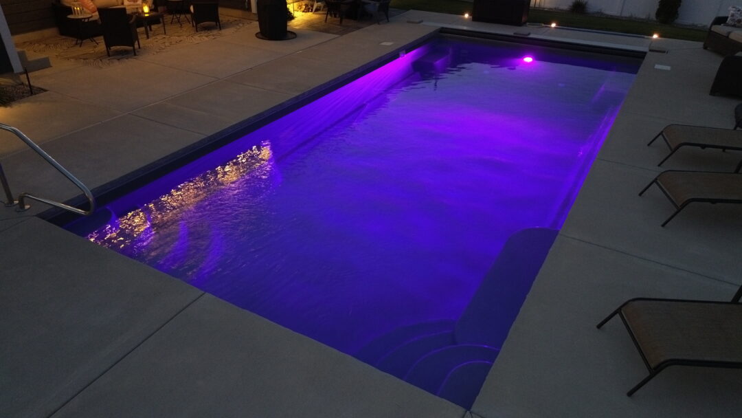 Landscape photo of an inground fiberglass pool with a purple light illuminating the pool. The pool is surrounded by gray concrete, atop which sit multiple lounge chairs surrounding the pool. On the far left side of the image is a handrail into the pool's shallow end cut off by the edge of the image. 