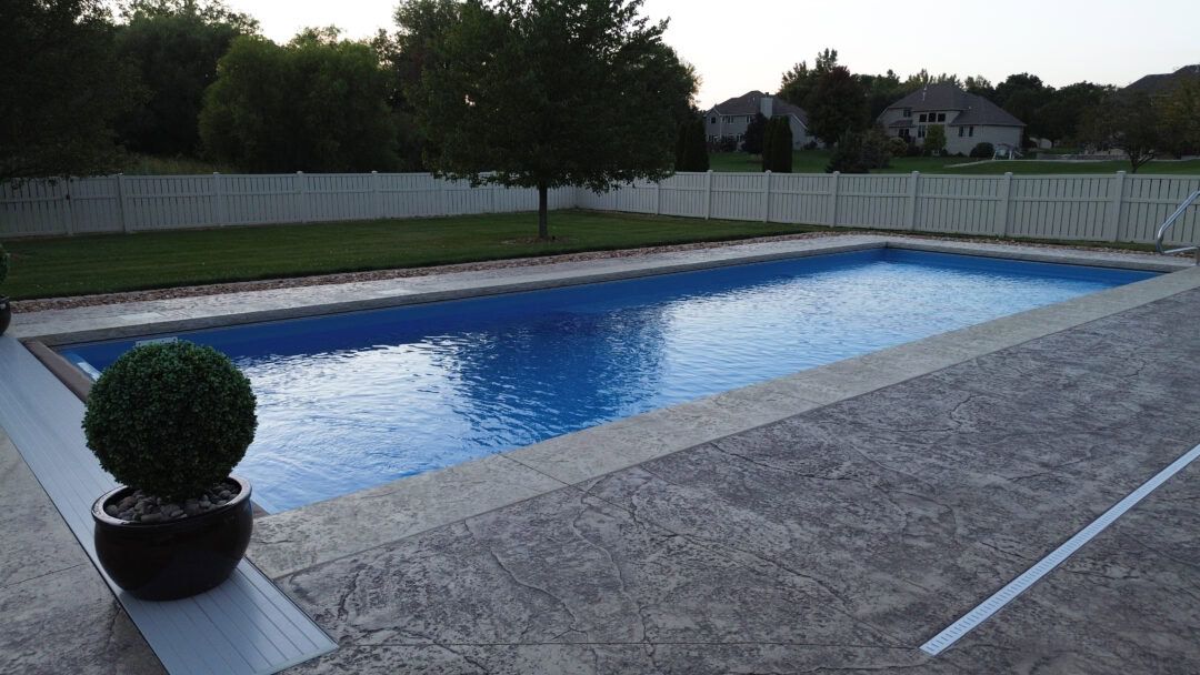 Landscape photo of a rectangular inground pool surrounded by a stamped concrete patio. The pool stretches the entire length of the image across the middle. Grass and two trees are behind the pool, and the yard is surrounded by a white fence. Beyond the fence, additional trees can be seen on the left, and more houses on the right.