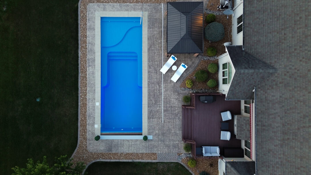 Aerial landscape photo of a portion of a backyard. In the center of the image is a stamped concrete patio within which sits a rectangular inground pool. Left of the patio is dark green grass. Right of the pool is a wood deck connected to the back of a house. A pergola roof sits off the right-side shallow end corner of the pool.