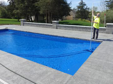 Landscape photo of a woman vacuuming the tanning ledge of a blue inground swimming pool. She is standing on the far right side of the image atop a gray stamped concrete patio. Behind her, parallel to the pools length is a retaining wall, separating the concrete from grasss behind it. Large trees and houes behind them can be seen in the background.