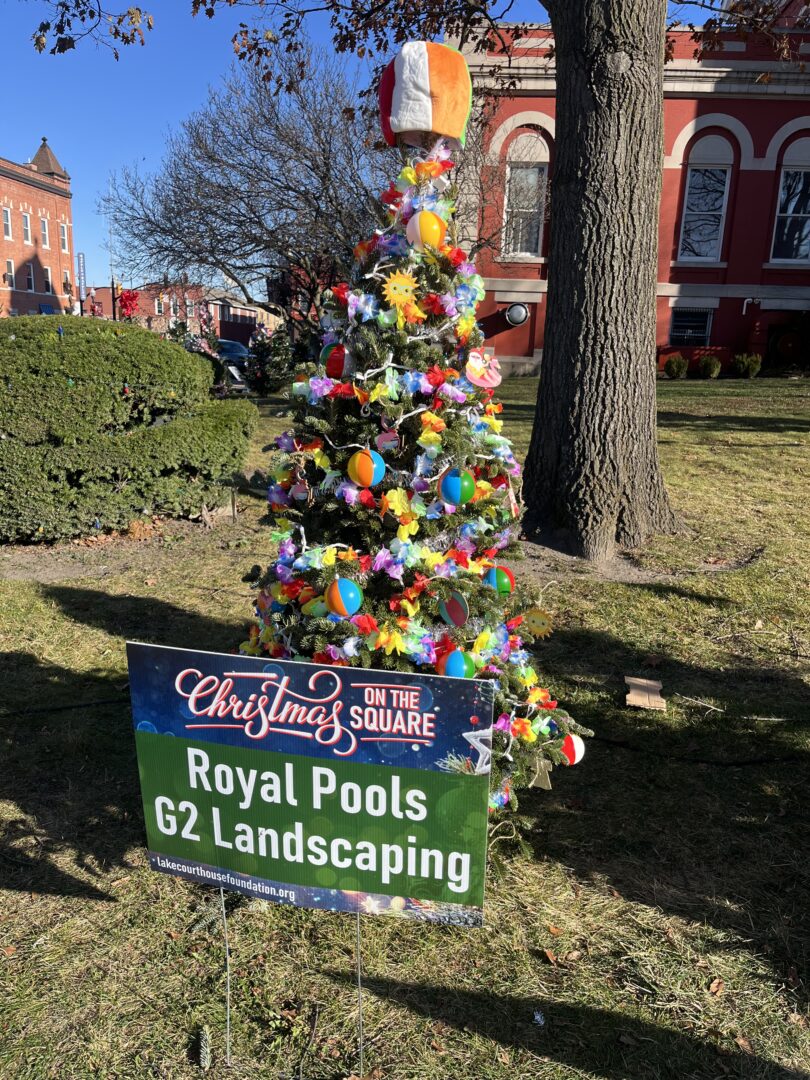 Portrait photo of a colorfully decorated Christmas tree in front of a tree and bush on the lawn in front of a red building. A sign in front of the Christmas tree says "Christmas ON THE SQUARE Royal Pools G2 Landscaping lakecourthousefoundation.org" Shadows can be seen going across the sign and tree. 