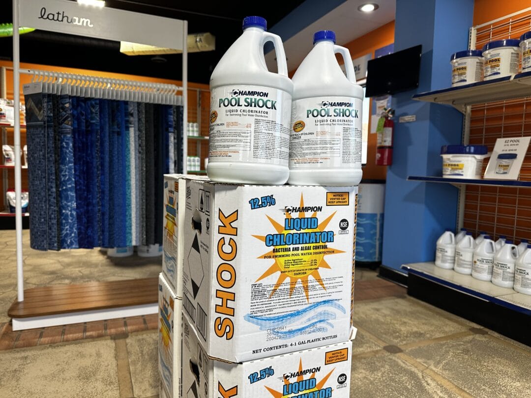 Landscape photo of a case of CHAMPION LIQUID CHLORINATOR SHOCK with two loose gallons stacked atop the case. Behind this display is a display of liners hanging.