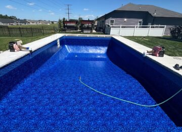 Landscape photo of an vinyl liner pool being filled with water via a garden hose sitting in the pool, spewing water into the deep end. The pool is surrounded by a concrete patio. On both sides of the pool's length are vacuums placed between the liner and walls. Grass surrounds the bpatio, and a fence can be seen in the background, behind which sits another house. More houses, trees, and blue sky can be seen in the background.