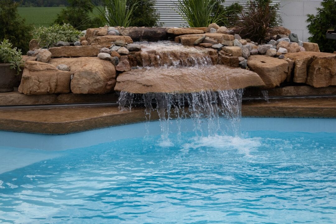 Image of a brown rock formation spilling over water into a light-blue inground swimming pool. A white house and shrubs can be seen in the background behind the pool.