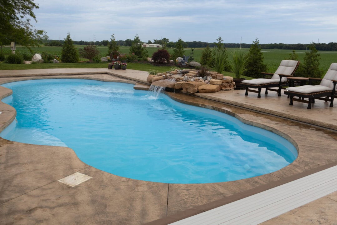 Landscape photo of a light blue, freeform inground pool surrounded by a tan stamped concrete patio. On the right side of the pool/image are two black-and-white lounge chairs. Next to the chairs, connected to the pool, is a rock formation waterfall. Lining the horizon and top quarter of the image are small evergreen trees in front of an empty field in the background. The sky can be seen above.